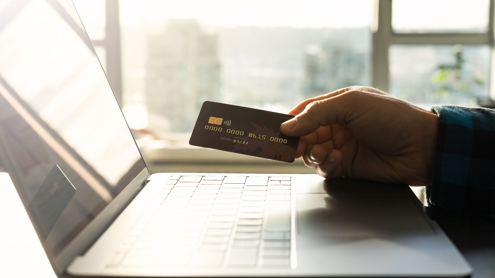 A shopper holding a payment card beside a laptop while comparing an online purchase